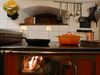 Ein alter Holzherd. Das Ofentürchen steht offen, im Ofen brennen ein paar Scheite. Auf der Kochplatte stehen eine schmiedeiserne Bratpfanne und ein oranger Bräter.