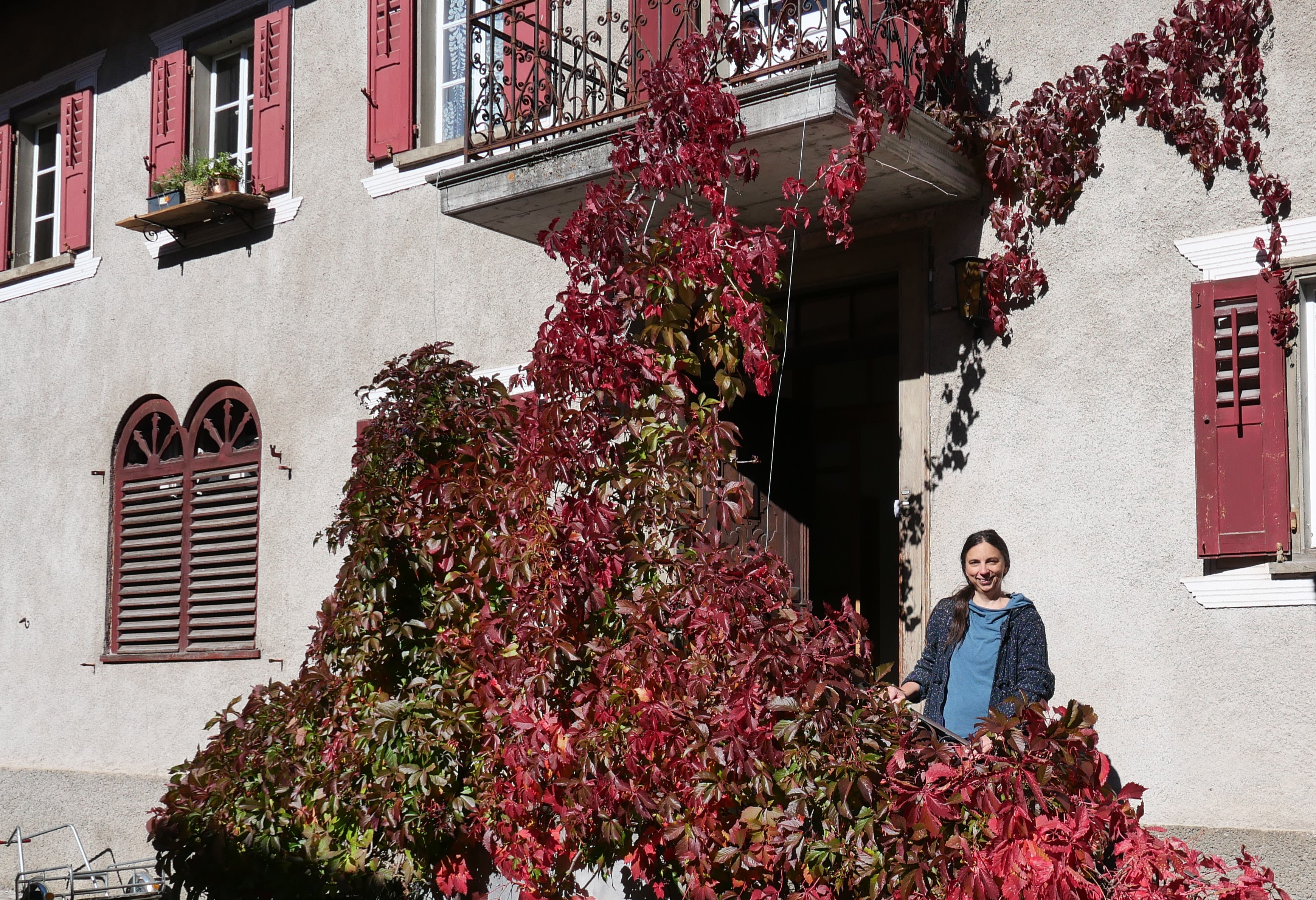 Eine Frau steht vor einem Haus und lächelt. Neben ihr eine herbstlich rot gefärbte Weinpflanze, dahinter die offene Haustür