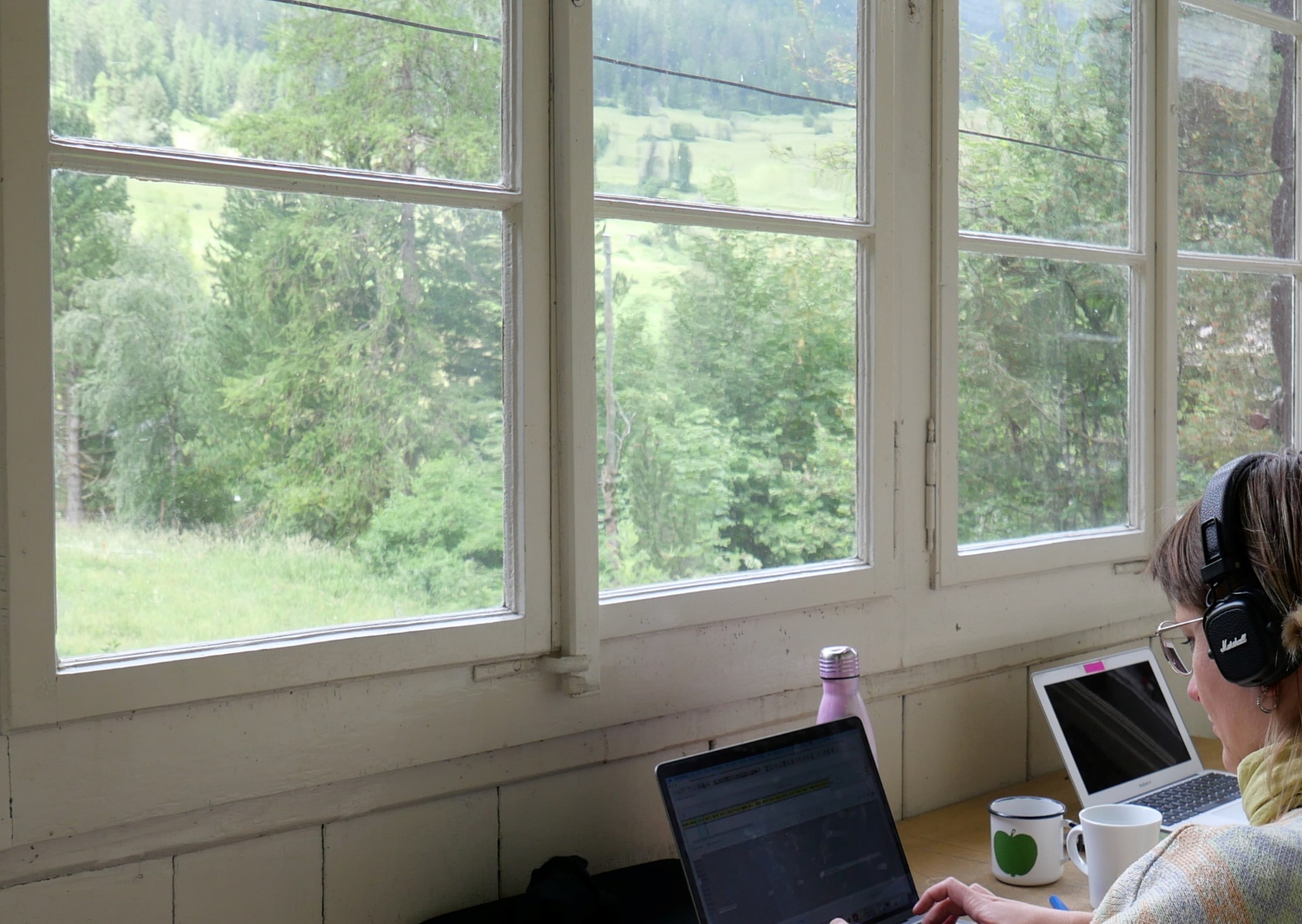 Eine junge Frau schreibt am Computer, durch die breiten Fenster ist eine grüne Berglandschaft zu sehen